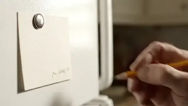 A man's hand writing a few lines on an index card held to a refrigerator door by a magnet